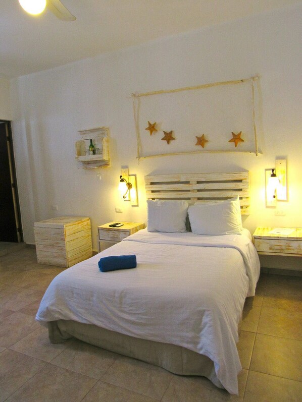 A neatly made bed with white linens, a wooden headboard, and a blue pillow. There are two bedside tables with lamps, a wooden chest of drawers, and a wall-mounted shelf with a bottle. - Coconut Hotel Boutique (Playa del Carmen)