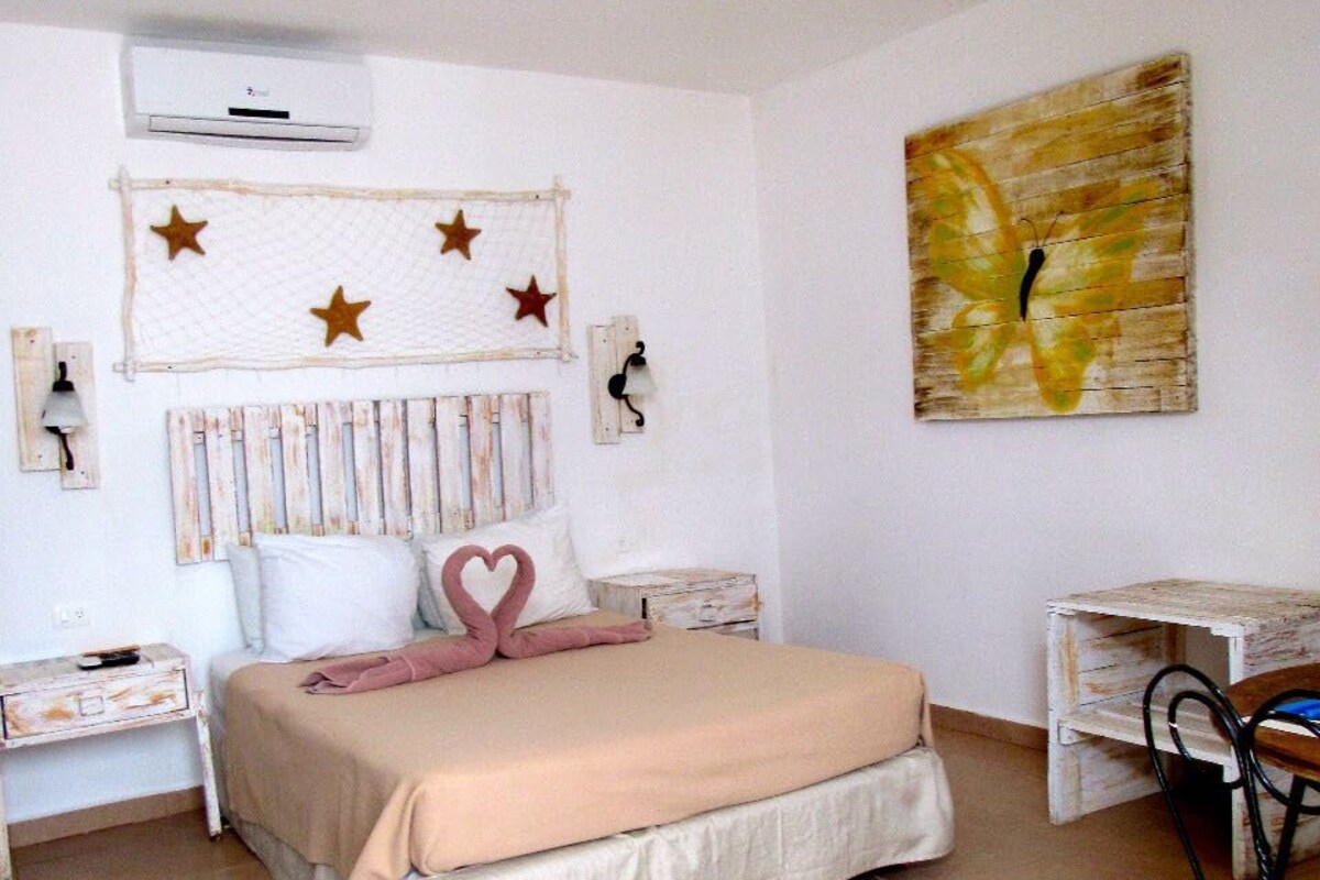 A bedroom with a bed, a ceiling fan, a wall-mounted air conditioner, a painting, and a small desk with a chair.