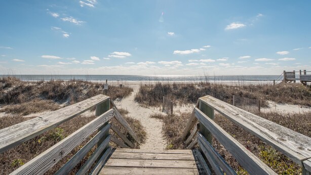 Une promenade en bois qui mène à une plage, avec un ciel dégagé et une vue sur l’océan à l’horizon.
