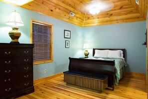 A bedroom with a wooden ceiling, a bed with a bench, a dresser, and a window with blinds.