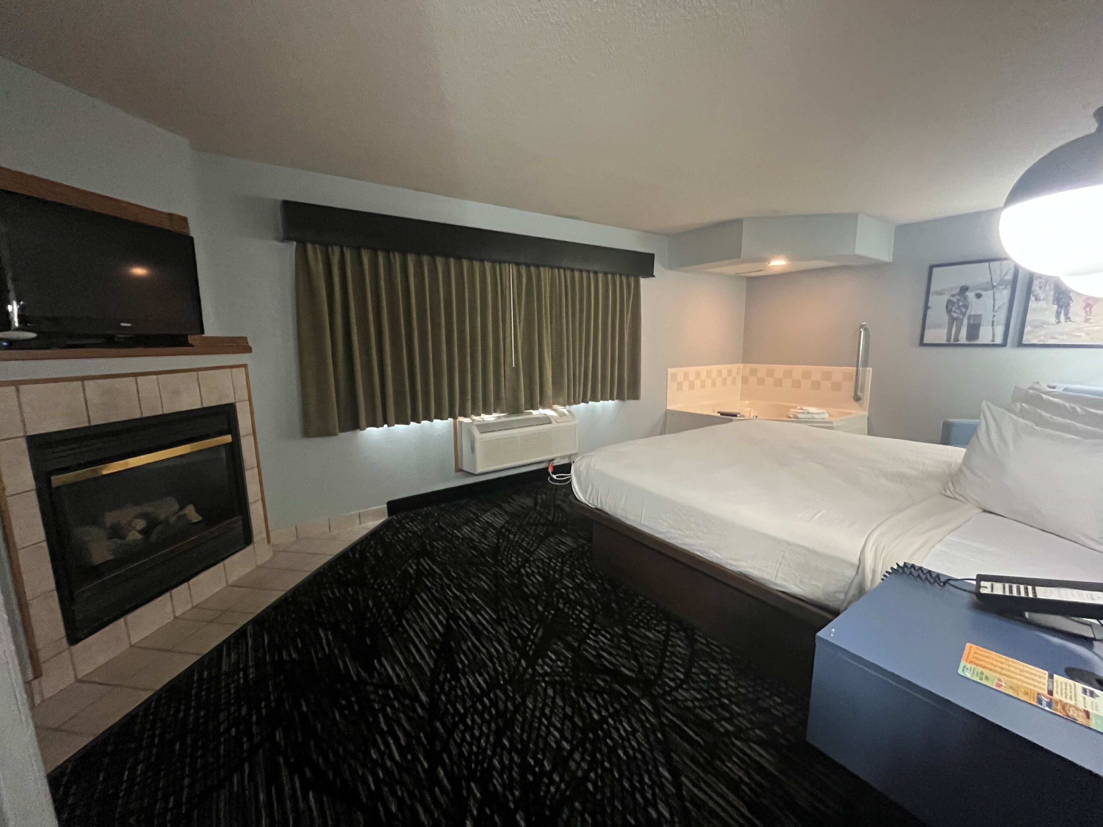 Quarto de hotel com lareira, cama com roupa de cama branca, escrivaninha com computador e janela com cortinas.