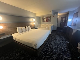 Habitación de hotel con una cama grande, lámparas de noche, cabecero, baño con bañera y un espejo.