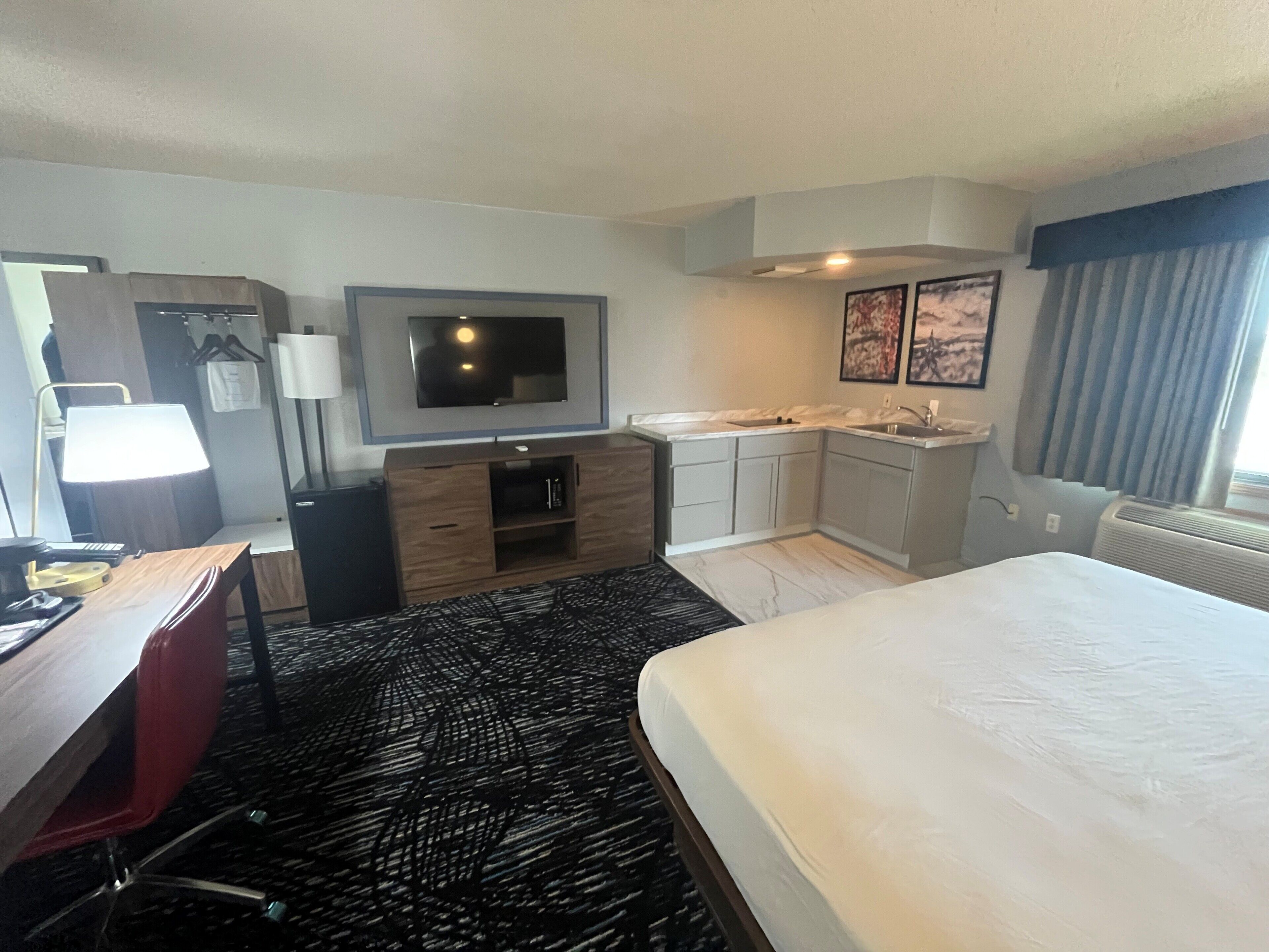Quarto de hotel com cama, escrivaninha, cadeira, televisão, bancada de cozinha e banheiro.
