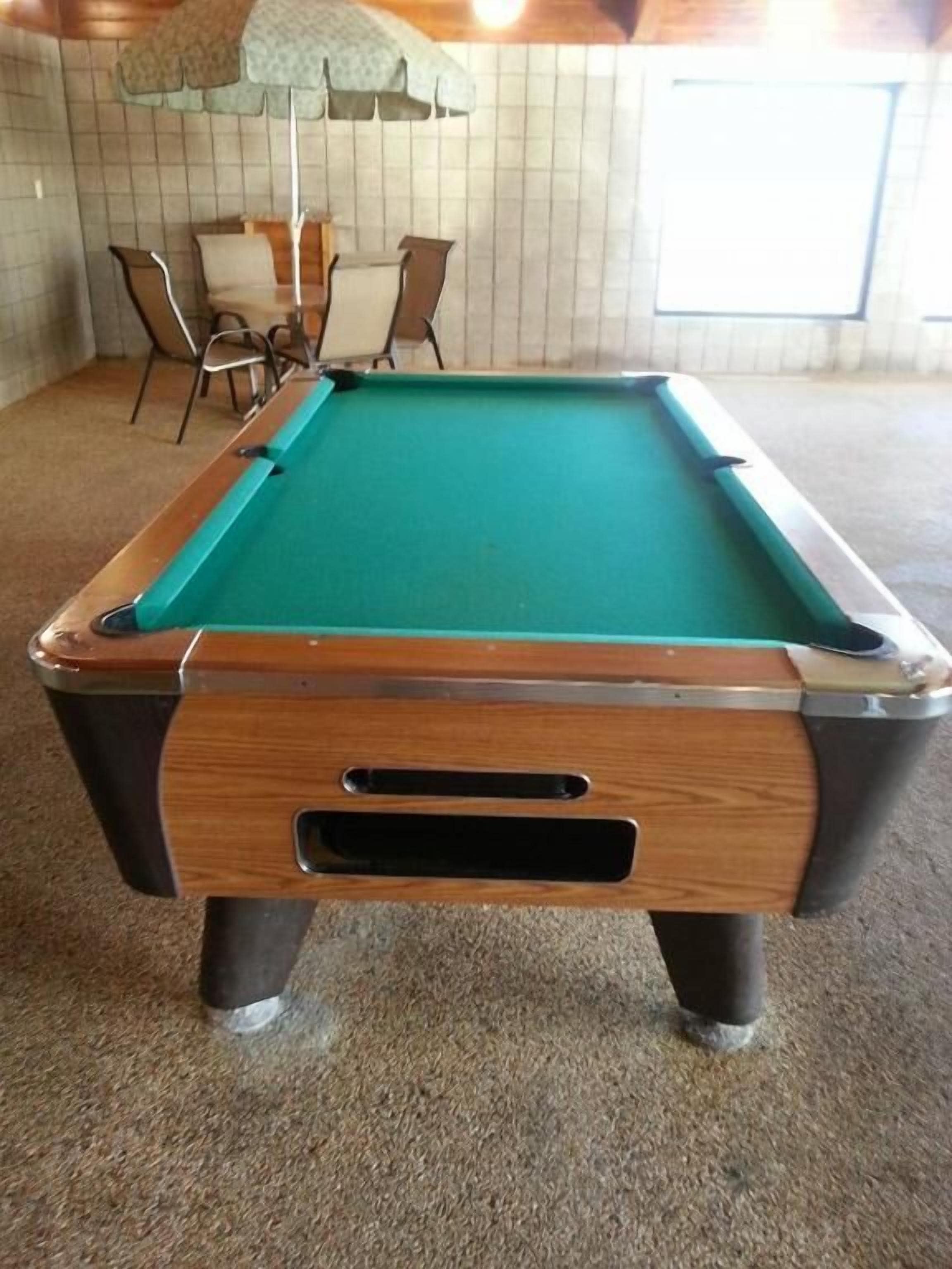 Una mesa de billar con marco de madera y superficie de fieltro verde, ubicada en una habitación con pared de azulejos y una ventana.
