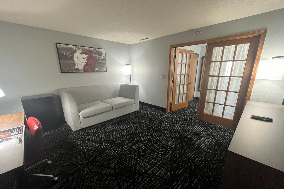 Habitación de hotel con sofá blanco, un cuadro en la pared, una lámpara, una puerta con paneles de vidrio y suelo alfombrado.