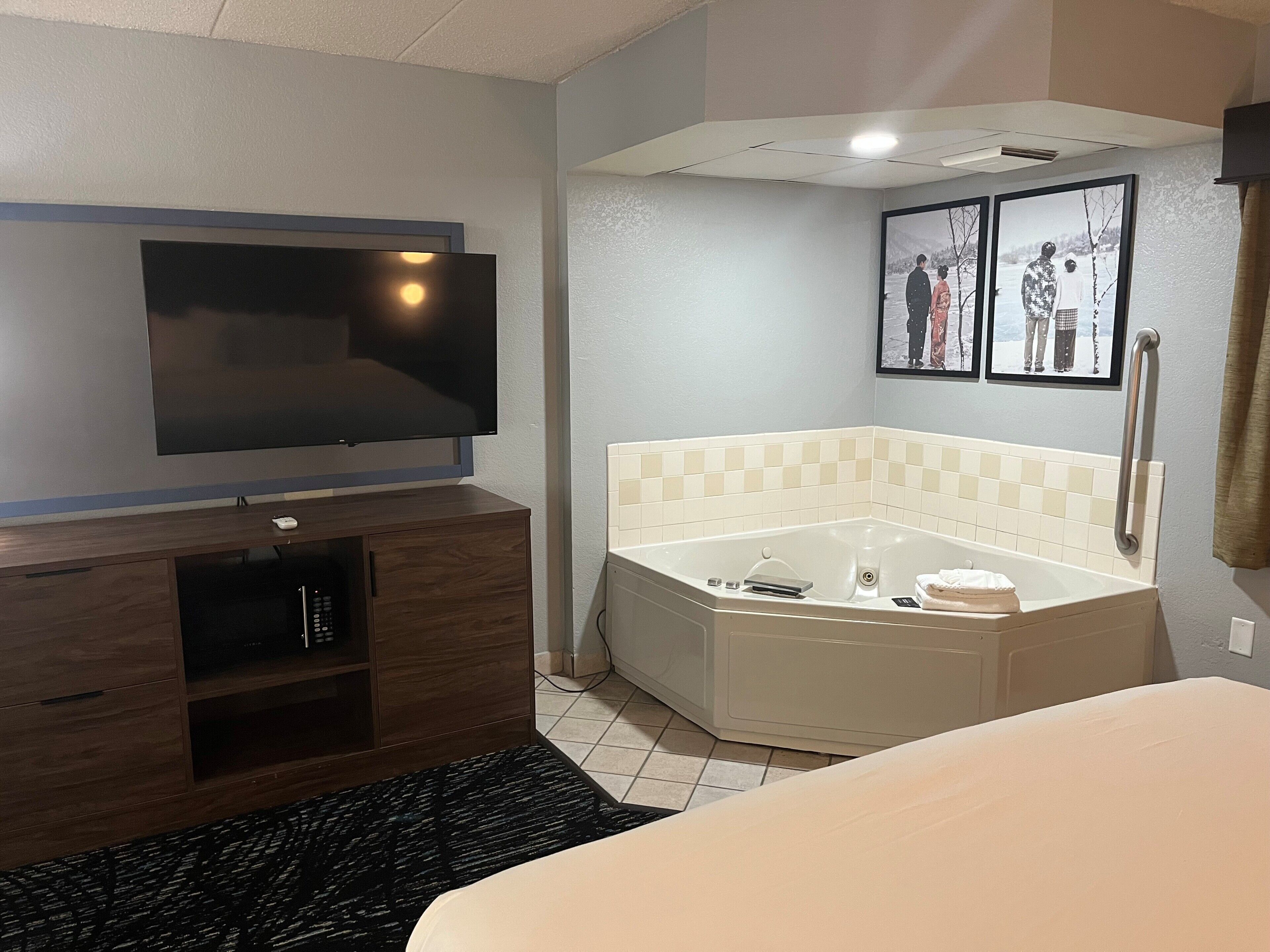 A hotel room with a flat-screen TV, a corner bathtub, a wooden cabinet, and a black rug.