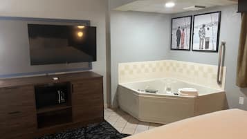 A hotel room with a flat-screen TV, a corner bathtub, a wooden cabinet, and a black rug.