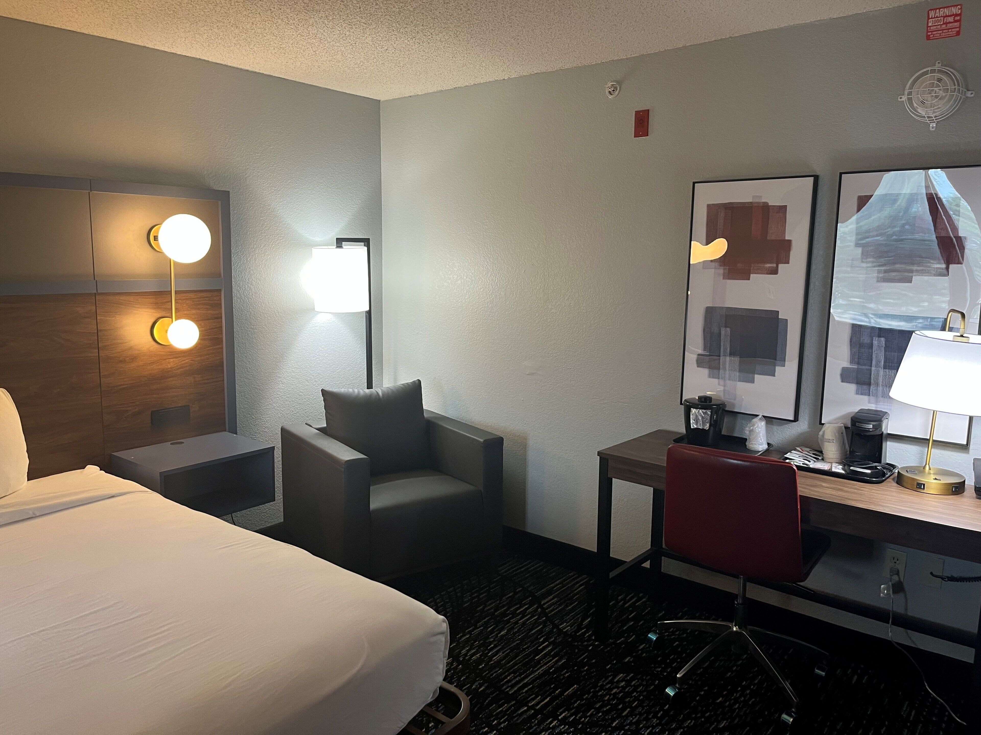 A hotel room with a bed, a desk, a chair, a lamp, and a wall-mounted mirror.