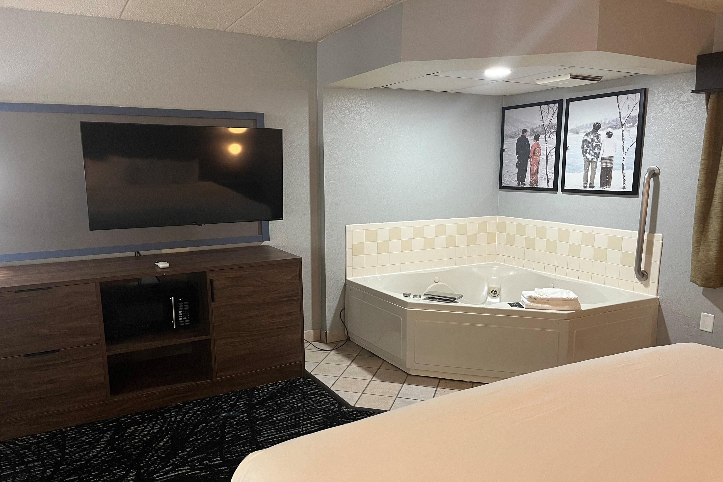 Habitación de hotel con televisor de pantalla plana montado en la pared, un mueble de madera con control remoto, una bañera en esquina con toalla y un libro, y dos cuadros en la pared.
