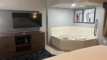 Habitación de hotel con televisor de pantalla plana montado en la pared, un mueble de madera con control remoto, una bañera en esquina con toalla y un libro, y dos cuadros en la pared.