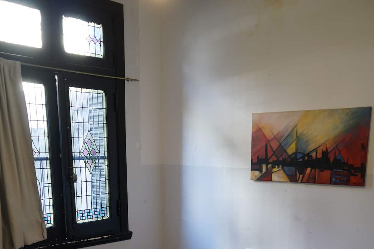 A hotel room with a large bed, a red bedspread, white pillows, a painting on the wall, and a window with a view of buildings.