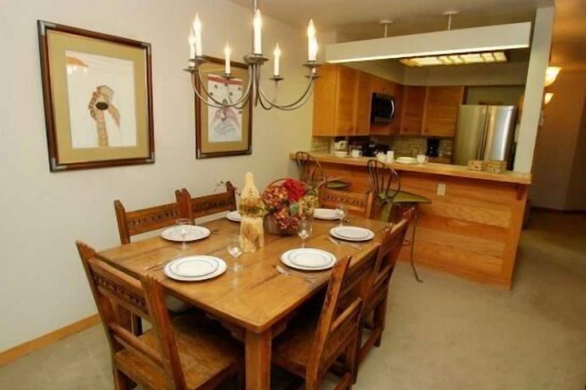 Un coin repas avec une table en bois dressée pour quatre personnes, entourée de chaises. Un lustre est suspendu au-dessus de la table et des œuvres d’art encadrées ornent les murs.
