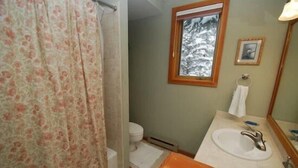 A bathroom with a shower curtain, a toilet, a sink with a mirror, and a window with a view of snow-covered trees. - Red Fox Town home 3 Bedroom Apartment by Key to the Rockies (Dillon)