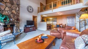 A spacious living area with a stone fireplace, leather sofas, a wooden coffee table, and a balcony with railings. - Snowdance Manor 2 Bedroom Holiday home by Key to the Rockies (Keystone)