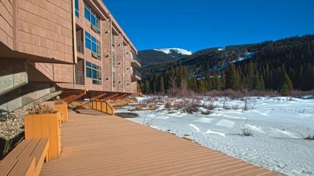 A wooden deck leading to a building with large windows, a snowy landscape, and a mountain in the background. - River Bank Lodge 1 Bedroom Apartment by Key to the Rockies (Keystone)