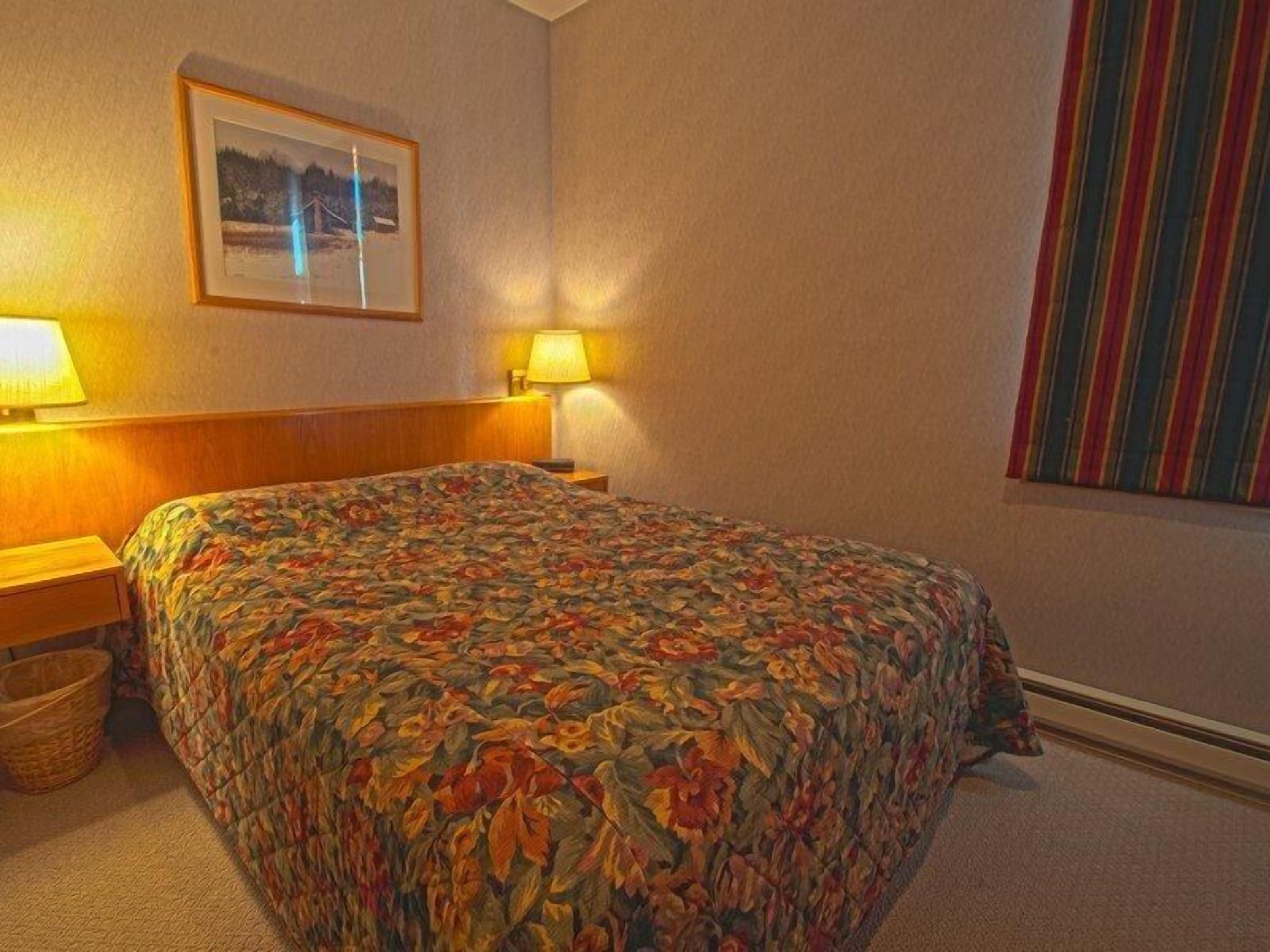 Une chambre d’hôtel avec un lit, une table de chevet, une lampe, un tableau encadré au mur et un rideau à rayures.