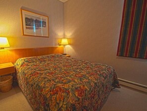 A hotel room with a bed, a nightstand, a lamp, a framed picture on the wall, and a striped curtain. - River Bank Lodge 1 Bedroom Apartment by Key to the Rockies (Keystone)