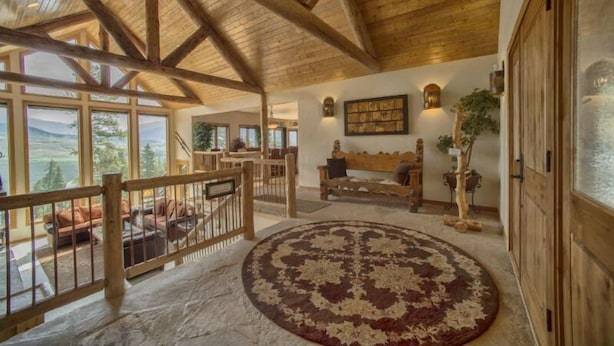 A spacious room with a high ceiling and wooden beams, a large window offering a view of the outdoors, a round rug, and a bench. - Retreat at Summerwood 5 Bedroom Apartment by Key to the Rockies (Dillon)