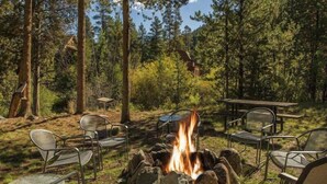 A campfire surrounded by metal chairs and tables in a forest setting.