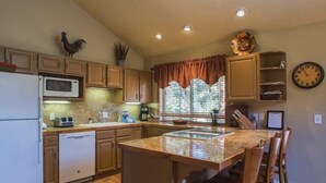 A kitchen with wooden cabinets, a microwave, a dishwasher, a sink, a window with curtains, a clock, and a bar stool.