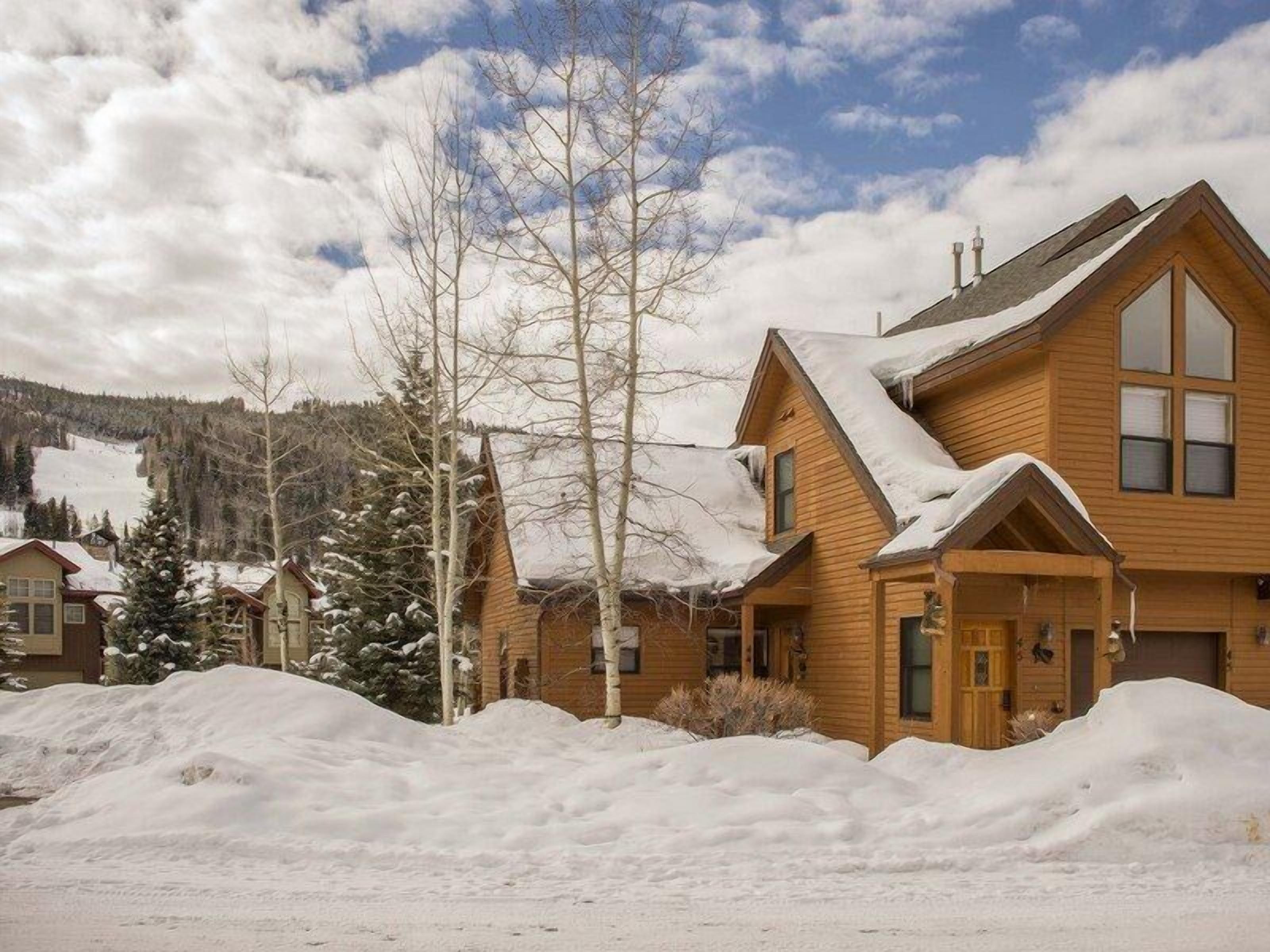 A wooden cabin with a steep roof, surrounded by snow-covered ground and trees.