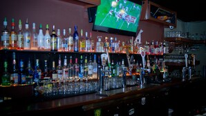 Un bar con scaffali pieni di bottiglie di liquore e bicchieri, un televisore che mostra una partita sportiva e una telecamera di sicurezza.