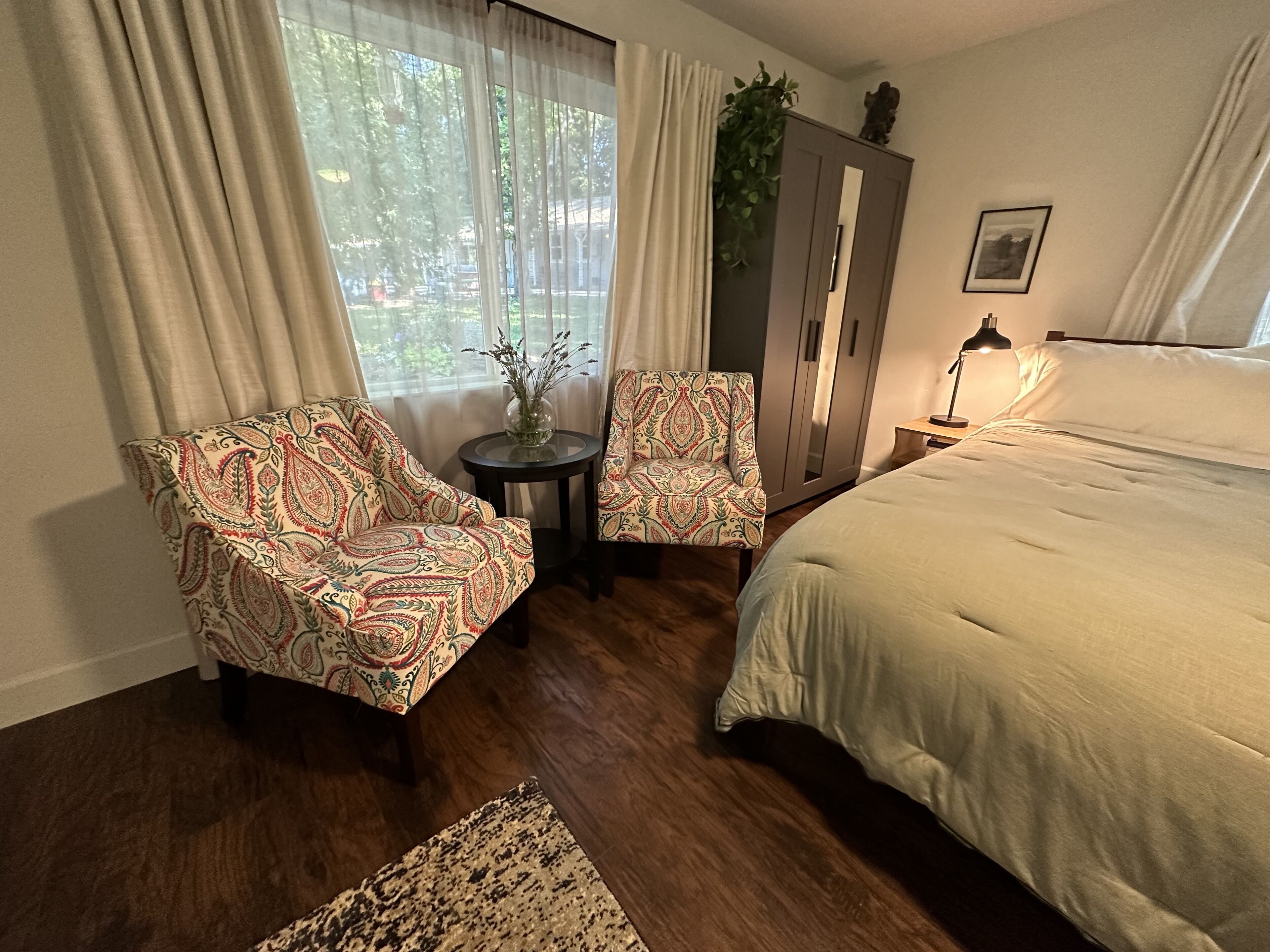 A bedroom with a bed, two patterned armchairs, a small table with a vase, a wardrobe, and a lamp.
