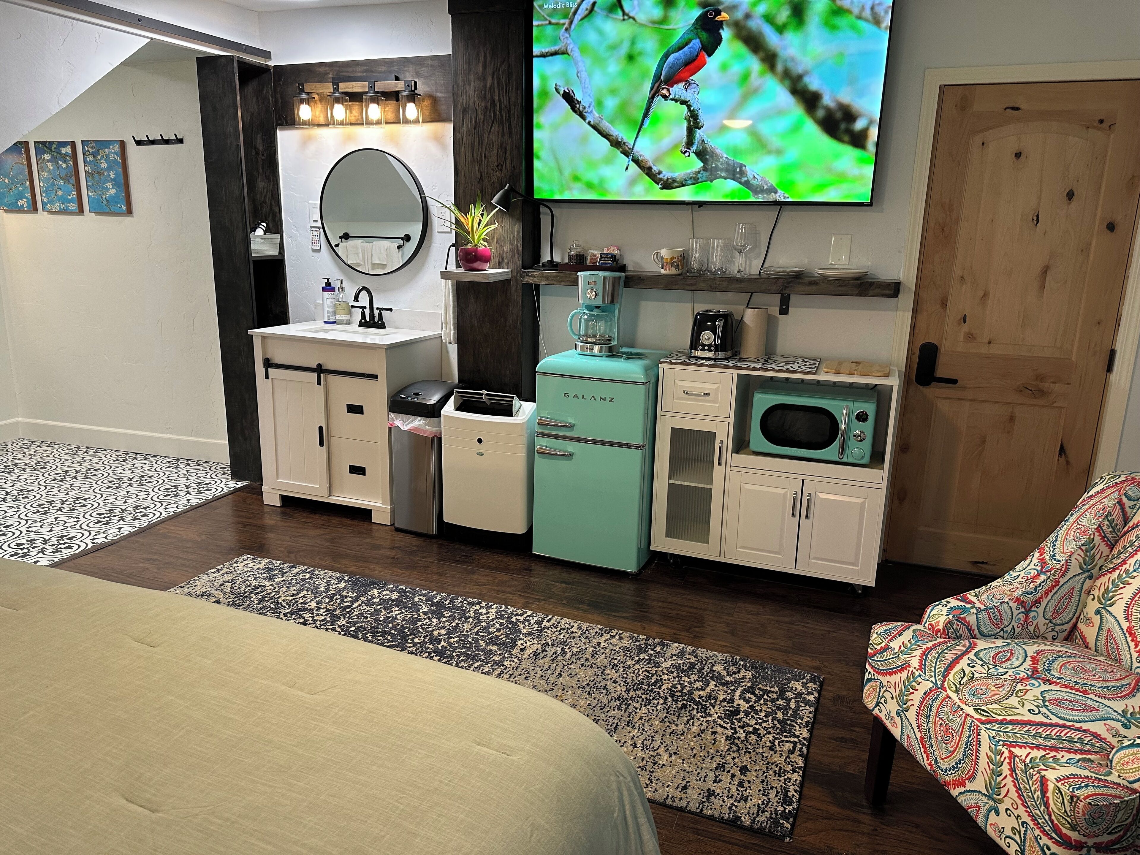 A compact bedroom with a bed, a vanity with a mirror, a small kitchenette with a fridge and microwave, and a television on the wall.