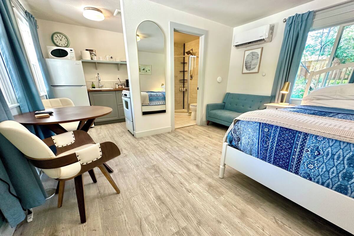 A compact room with a bed, a table, a chair, a kitchenette, and a bathroom.