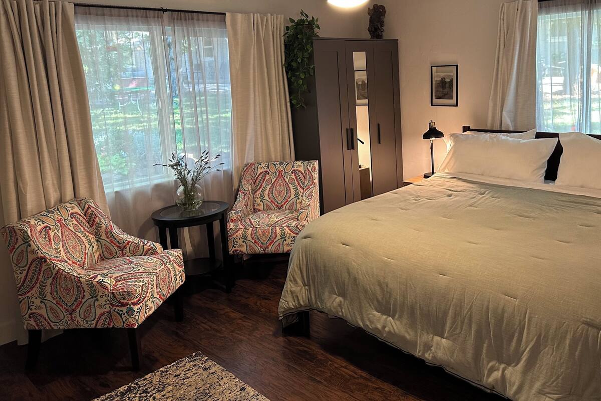 A bedroom with a large bed, two patterned armchairs, a small table with a vase, a wardrobe, and a window with curtains.