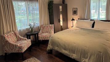 A bedroom with a large bed, two patterned armchairs, a small table with a vase, a wardrobe, and a window with curtains.