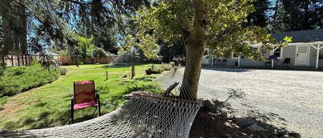 Una hamaca debajo de un árbol, una silla y un edificio con porche al fondo.
