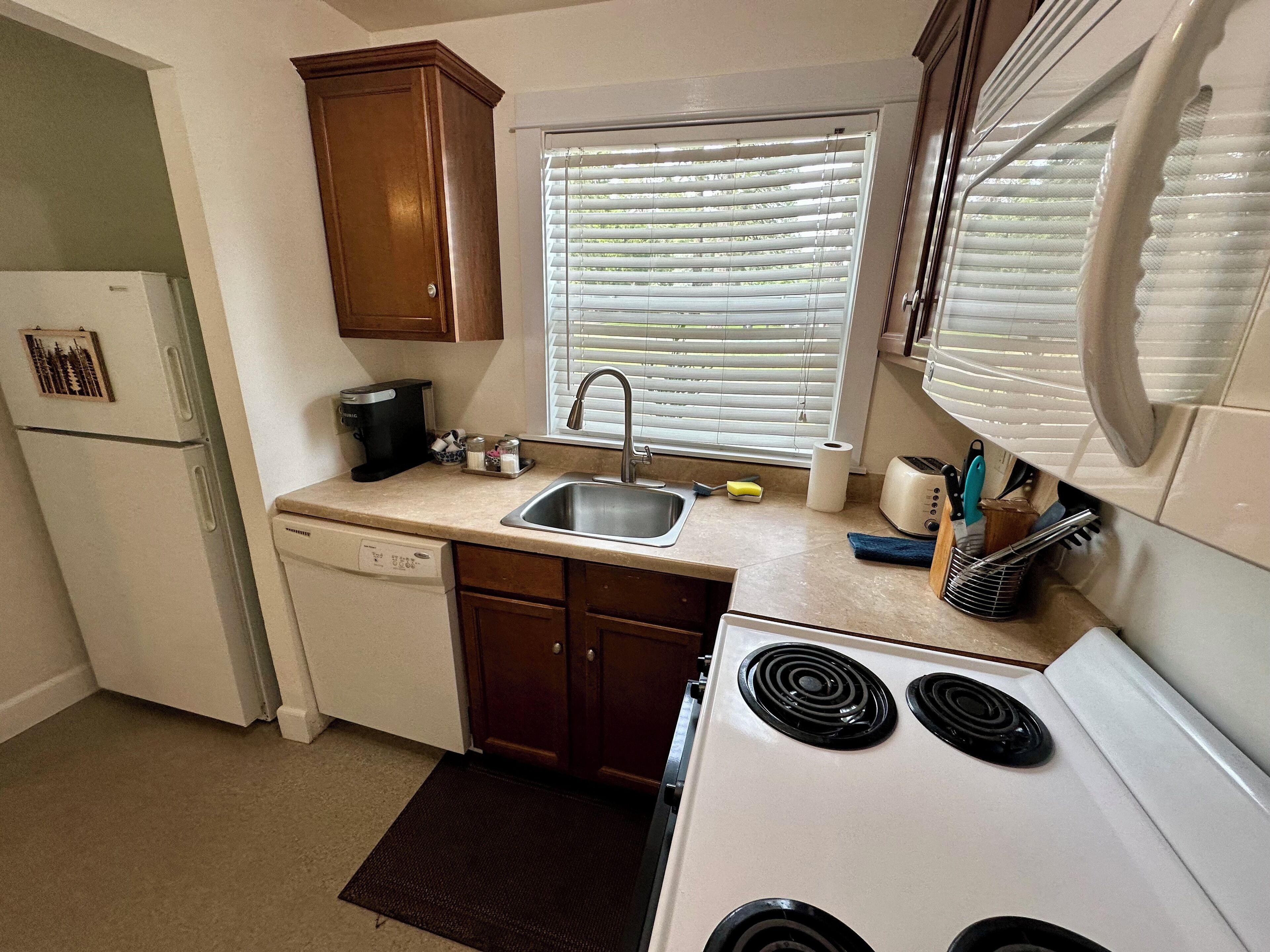 A compact kitchen with a refrigerator, dishwasher, stove, and a window with blinds.