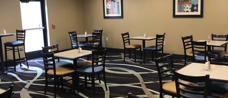 A hotel dining area with multiple tables and chairs, ceiling lights, and framed artwork on the walls.