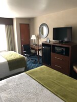 Habitación de hotel con dos camas, un escritorio con lámpara, televisión, minibar y un espejo en la pared.