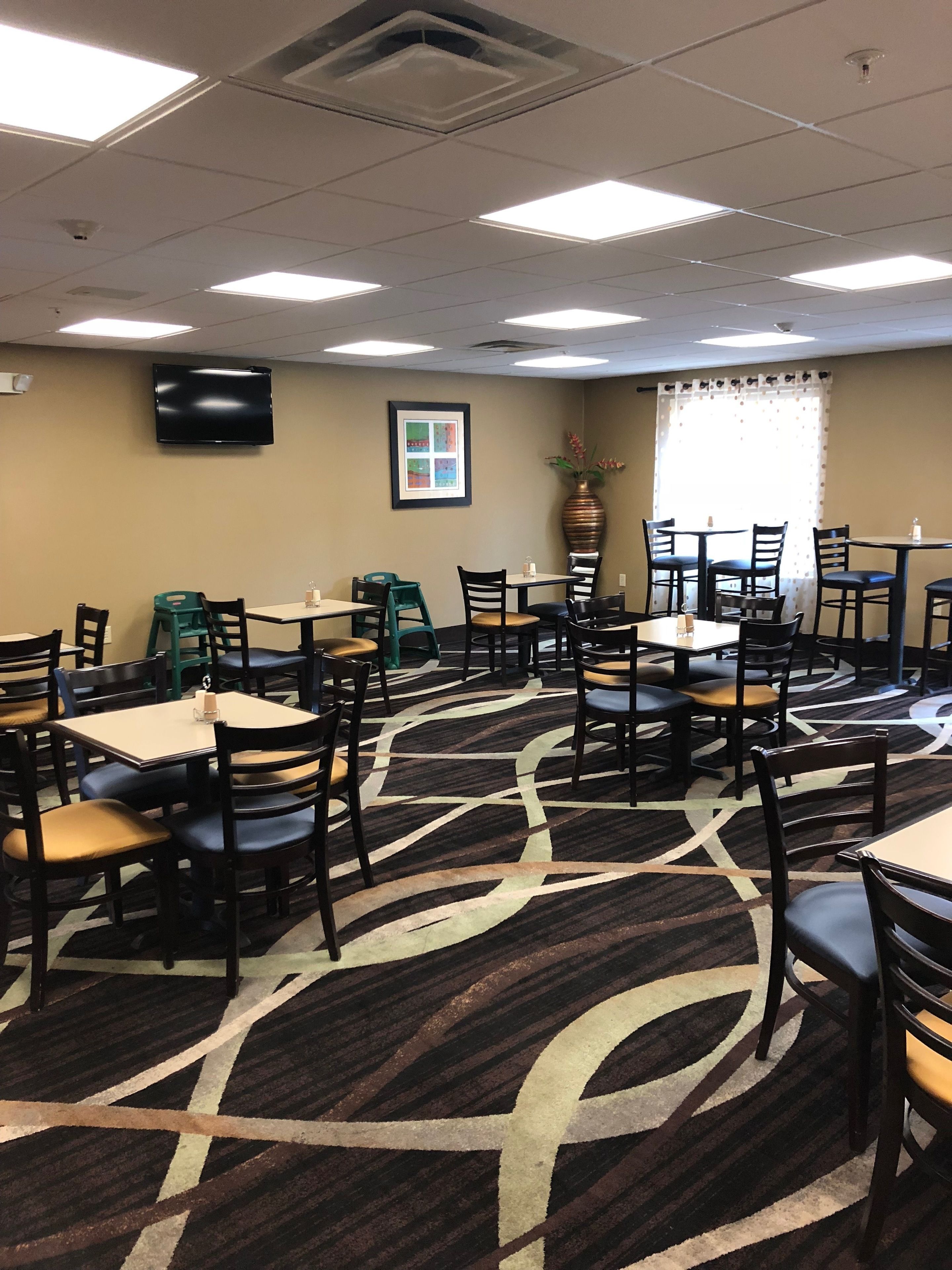 A hotel dining area with multiple tables and chairs, a flat-screen TV mounted on the wall, and a patterned carpet.