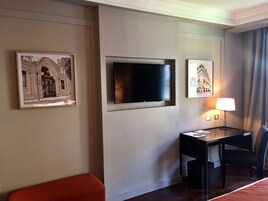 Habitación de hotel con televisor de pantalla plana en la pared, un escritorio con una silla y dos cuadros enmarcados.