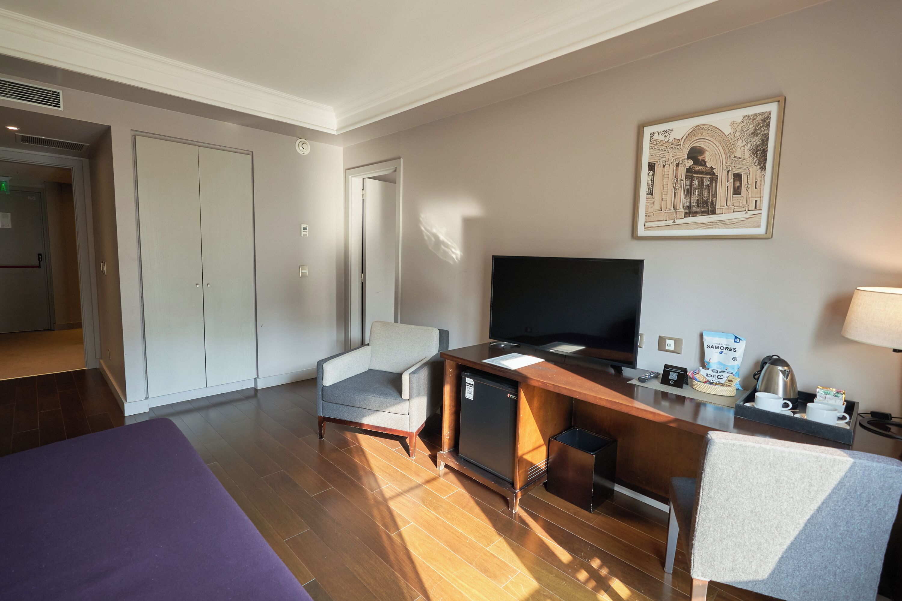 A hotel room with a bed, a desk with a television, a chair, a small refrigerator, and a coffee maker.