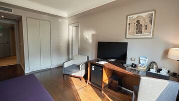A hotel room with a bed, a desk with a television, a chair, a small refrigerator, and a coffee maker.