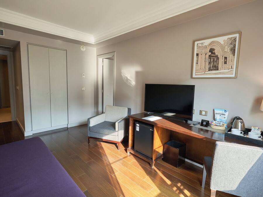 A hotel room with a bed, a desk with a television, a chair, a small refrigerator, and a coffee maker.