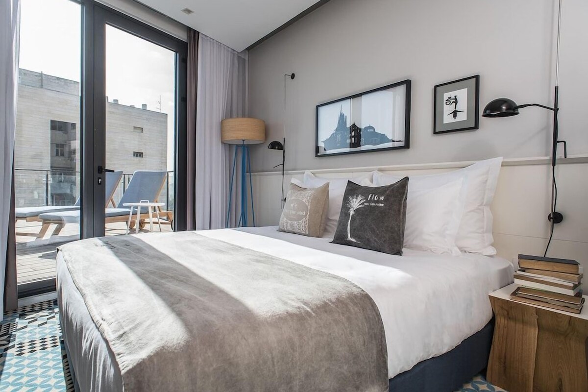 Habitación de hotel con una cama grande, vistas a la ciudad y un balcón con mobiliario exterior.