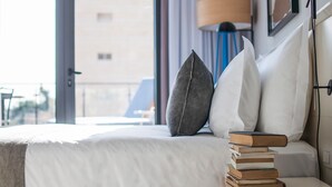 Een net opgemaakt bed met een witte dekbedovertrek en kussens, een stapel boeken op een houten nachtkastje, en uitzicht buiten door een groot raam.