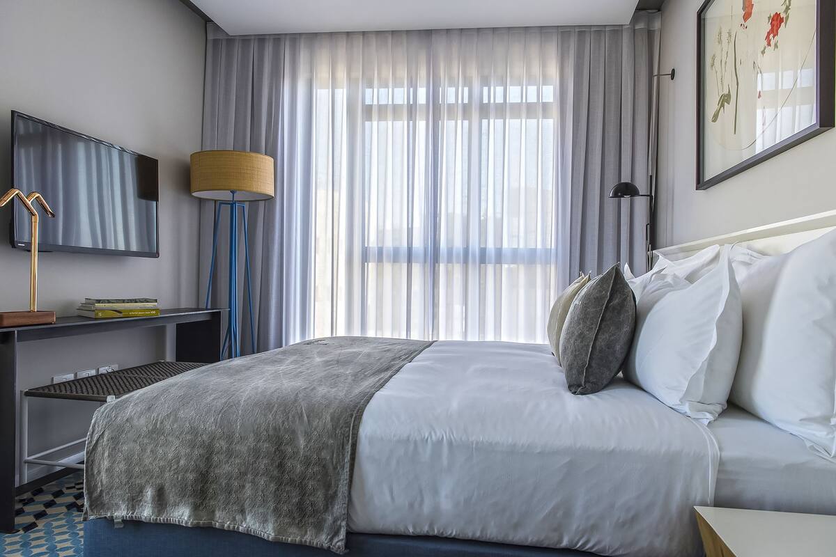 Habitación de hotel moderna con una cama grande, un escritorio con televisor de pantalla plana, una lámpara y un ventanal con cortinas translúcidas.