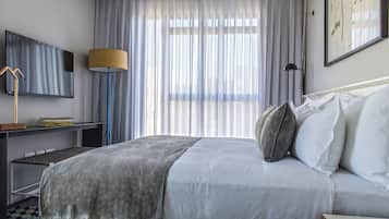 Une chambre d’hôtel moderne dotée d’un grand lit, d’un bureau avec un téléviseur à écran plat, d’une lampe et d’une grande fenêtre ornée de rideaux légers.