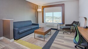 A modern hotel room with a blue sofa, a wooden coffee table, a desk with a chair, a large window with a view of the city, and a floor lamp. - Holiday Inn Express & Suites Covington by IHG (Covington)