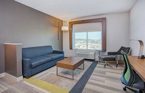 A modern hotel room with a blue sofa, a wooden coffee table, a desk with a chair, a large window with a view of the city, and a floor lamp. - Holiday Inn Express & Suites Covington by IHG (Covington)