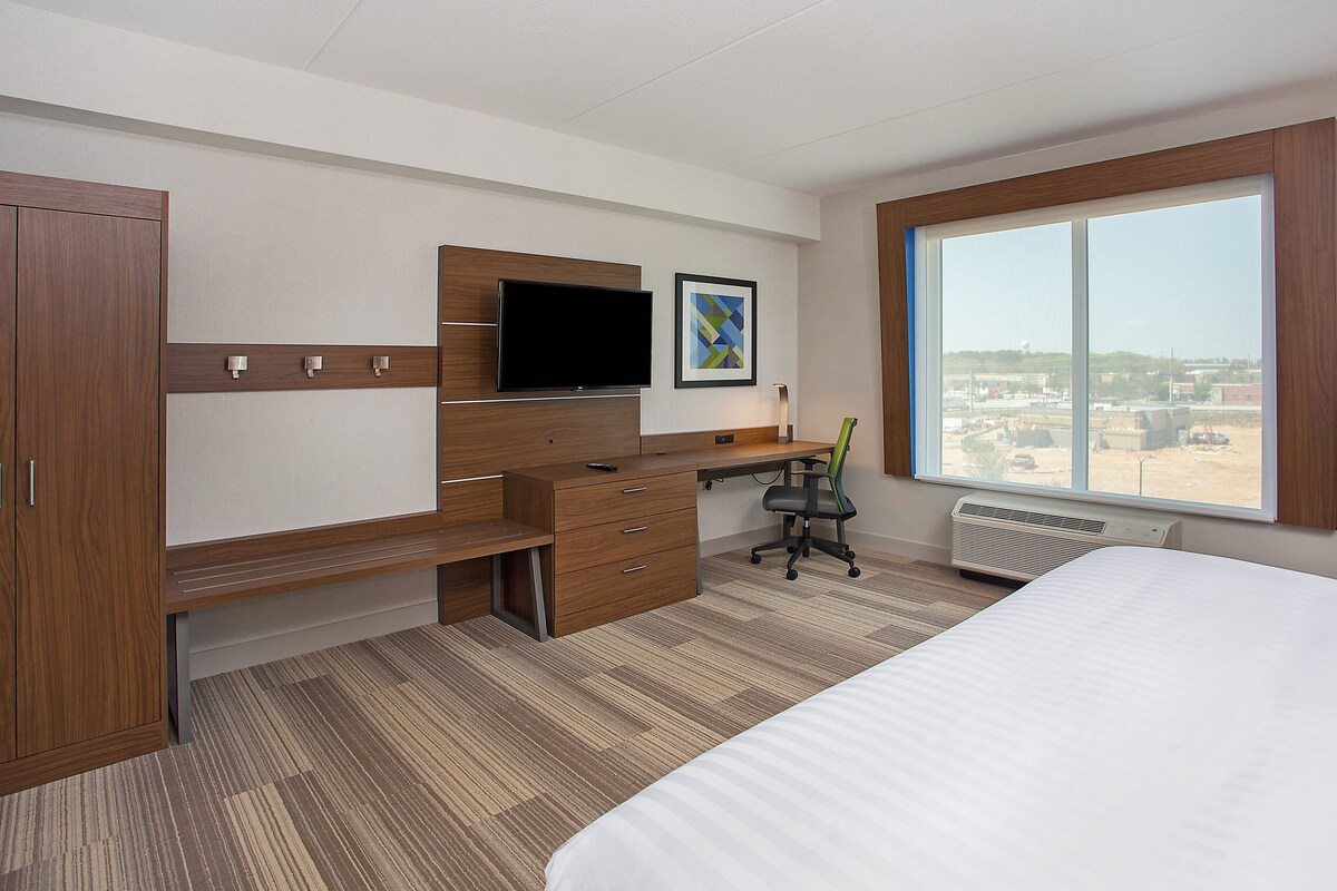 A hotel room with a bed, a desk with a flat-screen TV, a chair, a radiator, and a large window with a view.
