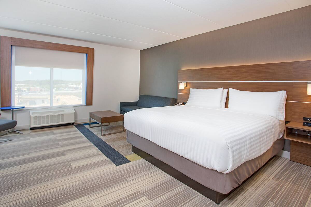 A modern hotel room with a large bed, a desk, a chair, and a view of the city through a large window.
