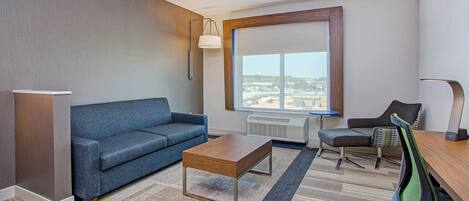 A modern hotel room with a sofa, a wooden coffee table, a desk with a chair, a large window with a view of the city, and a standing lamp.