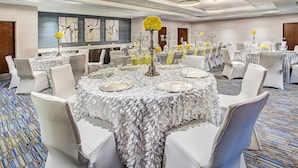 Salão de banquetes com mesas redondas preparadas para refeições, cadeiras brancas e centros de mesa decorativos.